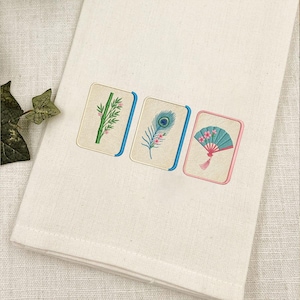 Puede incluir: Una toalla de tela color crema con tres parches rectangulares bordados. Los dise&ntilde;os muestran un tallo de bamb&uacute; verde, una pluma de pavo real azul y verde, y un abanico plegable color cerceta con flores rosas, delineados en azul o rosa.
