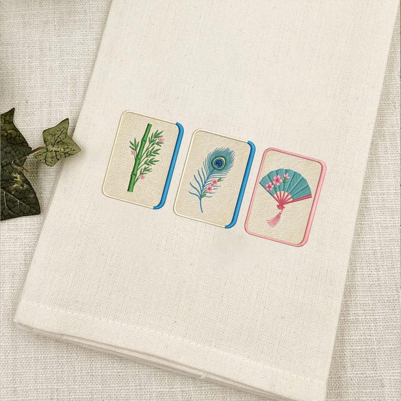 Puede incluir: Una toalla de tela color crema con tres parches rectangulares bordados. Los dise&ntilde;os muestran un tallo de bamb&uacute; verde, una pluma de pavo real azul y verde, y un abanico plegable color cerceta con flores rosas, delineados en azul o rosa.