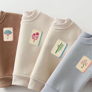 Puede incluir: Cuatro sudaderas de cuello redondo en marr&oacute;n, crema, beige y azul claro. Cada sudadera presenta un parche rectangular en el pecho que representa una ficha de mahjong con ilustraciones de un abanico, flores, bamb&uacute; y un &aacute;rbol.
