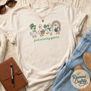 May include: Cream-colored t-shirt featuring a St. Patrick's Day design. The graphic shows four cartoon geese with festive accessories, a rainbow, and the text "just a lucky goose" in green. The shirt is paired with blue jeans and brown boots.