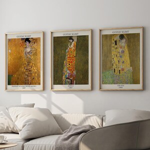 Puede incluir: Tres impresiones artísticas enmarcadas con obras de Gustav Klimt cuelgan sobre un sofá de color claro. Las impresiones tienen tonos dorados, verdes y amarillos. El sofá tiene cojines blancos y una manta gris. Una mesa auxiliar de madera y una silla moderna completan el salón.