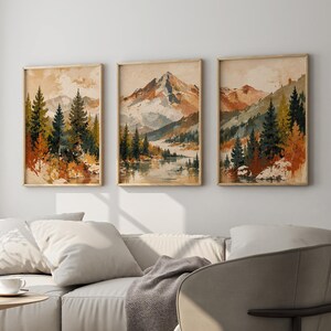 May include: Three vertical framed wall art prints featuring watercolour-style mountain landscapes with pine trees, rocky peaks, and calm water in shades of rust, forest green, cream, and brown, displayed in a row on a plain light-coloured wall.