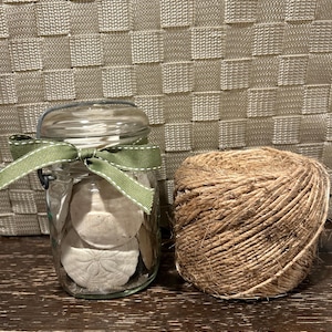 Puede incluir: Un tarro de cristal transparente lleno de dólares de arena, atado con una cinta verde y una bola de cuerda. El tarro está sobre una superficie de madera oscura, con una cesta tejida en el fondo.