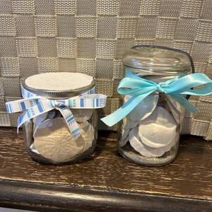 Puede incluir: Dos tarros de vidrio llenos de dólares de arena y arena. Un tarro tiene una tapa blanca y una cinta azul a rayas. El otro tarro tiene una tapa transparente y una cinta azul claro atada alrededor del cuello.