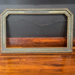 May include: A rectangular picture frame with clipped corners, featuring a distressed blue and gold finish. The frame includes ornate floral carvings at the top center and decorative scrollwork at the bottom corners, with a thin wire attached to the back.