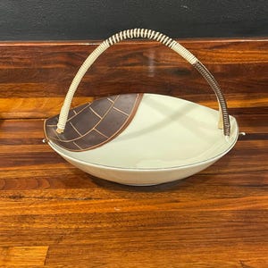 May include: Oval-shaped serving dish with a woven handle. The dish is off-white with a brown geometric design on one side. The handle is a combination of woven beige and brown materials.