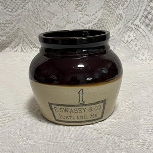 May include: A vintage stoneware crock with a dark brown top and a tan base. The crock has the number "1" and the text "E. SWASEY & CO. PORTLAND, ME" printed on the front. The crock is likely used for storage.