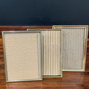 May include: Three gold-colored picture frames of varying sizes. The frames have a simple, elegant design with a thin border. The frames are empty, with cardboard backing visible. The frames are arranged on a wooden surface.