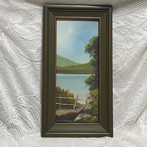 May include: A framed landscape painting depicting a serene lake scene. The artwork features a wooden fence, lush green trees, and a mountain range under a blue sky. The frame is a dark green color.