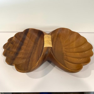 May include: A wooden serving dish with a unique design. The dish is made of dark brown wood and features two shell-shaped compartments connected by a woven rattan handle. The shell-shaped bowls have carved ridges.