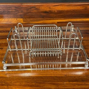 May include: A rectangular clear acrylic tray featuring a polished chrome metal wire rack system. The metal structure includes a central grid section flanked by four upright U-shaped dividers, all supported by small spherical chrome feet at each corner.