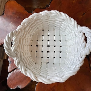 May include: A white ceramic basket with a woven design. The basket has a rectangular shape with rounded corners and two rope-like handles. The basket is sitting on a wooden surface.