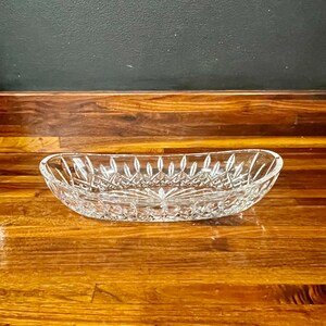 May include: An oval-shaped clear glass dish featuring a detailed cut crystal pattern. The design includes a series of vertical facets along the rim and a cross-hatch diamond motif covering the exterior surface, with a starburst pattern at the base.