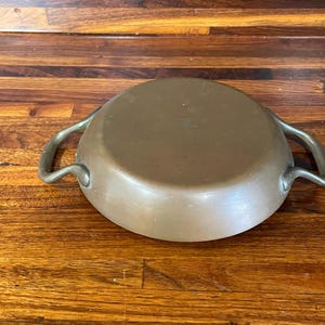 May include: A copper-colored, round cooking pot with two curved metal handles. The pot is sitting on a wooden surface with a warm, brown tone. The pot's interior is not visible.