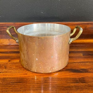 May include: A round copper cooking pot with a metallic silver interior lining. The exterior surface of the pot shows a weathered patina with visible spots and discoloration. Two curved brass handles are attached to opposite sides.