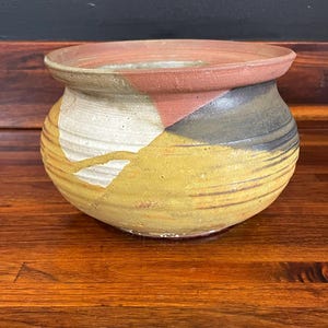 May include: A handcrafted ceramic bowl with a wide, flat rim. The bowl features a color-blocked design with sections of white, yellow, brown, and black. The pottery has a rustic, textured appearance, suggesting it is handmade.