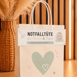 Könnte beinhalten: Eine hellbeige Papiertüte mit weißen Henkeln, verziert mit einem großen salbeigrünen Herz und kleineren rosa Herzen. Ein rechteckiges Etikett ist angebracht, das den Text NOTFALLTÜTE für Mamas & Papas FÜR ALLE FÄLLE. FÜR EUCH. sowie eine Grafik eines kleinen Beutels zeigt.