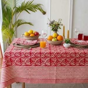 May include: A rectangular dining table covered with a red and white patterned tablecloth featuring a repeating leaf motif and striped border. The table is set with two bowls of fresh fruit, glasses of orange juice, and folded napkins on woven mats.
