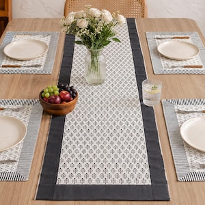 Puede incluir: Un juego de mesa que incluye un camino de mesa largo y cuatro manteles individuales rectangulares a juego, decorados con un patrón de árboles en blanco y negro y bordes negros sólidos. La mesa está puesta con cuatro platos blancos, cubiertos, un vaso y un frutero.