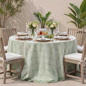 May include: A round dining table covered with a sage green and white patterned tablecloth, surrounded by four wooden chairs with white cushions. The table is set with woven placemats, white plates, napkins, and two floral centerpieces.