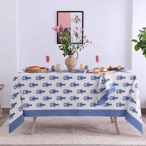 May include: A rectangular tablecloth featuring a repeating pattern of blue lobsters on a white background with a solid blue border. The fabric drapes over a table set with a central ceramic vase of pink flowers, glassware, and small food items.