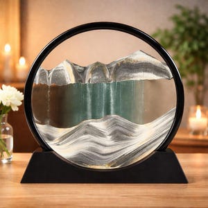 Peut inclure: Une œuvre d'art décorative en sable avec un cadre et une base circulaires noirs. À l'intérieur, des couches de sable créent une scène de montagne et d'eau avec des nuances de gris, d'or et de vert. L'œuvre est posée sur une surface en bois, avec des bougies et des fleurs en arrière-plan.