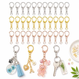 May include: A collection of metal keychain hardware in silver, gold, and rose gold tones, featuring lobster clasps and chains. Four finished keychains are displayed, including charms like a blue tassel, a heart, a bear, and a fern-filled resin piece.