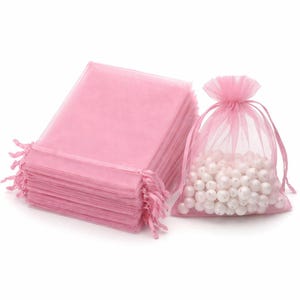 May include: A collection of pink organza bags, one filled with white pearls. The bags are a light, translucent pink. The filled bag is tied with a pink drawstring. The stack of bags has fringed edges.