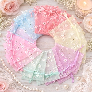 Puede incluir: Una colección de bolsas transparentes con cordón, dispuestas en círculo. Cada bolsa presenta un delicado estampado floral blanco y una cinta a juego. Los colores incluyen rosa, azul, amarillo y verde. Ideales para regalos.