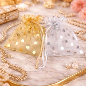 May include: Two sheer gift bags, one gold and one white, are tied with matching ribbons. The bags are decorated with large, metallic polka dots. The background includes jewellery and decorative elements, suggesting a theme of gifts or accessories.