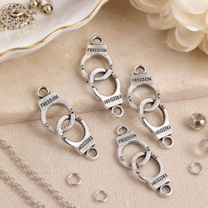 May include: Four silver-toned metal charms shaped like linked handcuffs, each featuring the word FREEDOM embossed on the surface. These jewelry components include small loops at the top and bottom for attachment, presented with a silver chain.