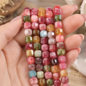 May include: A strand of faceted, multi-colored gemstone beads in shades of pink, red, green, yellow, and blue. The beads are square-shaped with rounded edges and a polished finish, strung together on a thin cord to form a continuous, vibrant line.