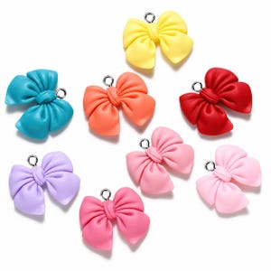 Peut inclure: Un ensemble de huit breloques en forme de nœud de différentes couleurs unies, notamment jaune, bleu sarcelle, orange, rouge, lavande, rose vif, rose clair et rose pâle. Chaque breloque possède un petit anneau en métal argenté au sommet.