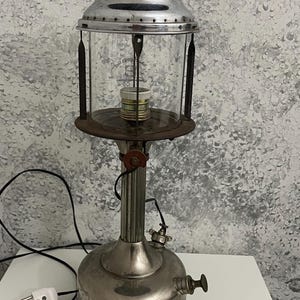 Könnte beinhalten: Vintage silberfarbene Laternenlampe mit einem transparenten Glasgehäuse. Die Lampe hat einen runden Sockel, einen zylindrischen Körper und eine Oberseite mit Lüftungslöchern. Ein schwarzes Netzkabel mit weißem Stecker geht von der Basis aus.