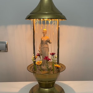 Può includere: Una lampada vintage in ottone con una statua classica all'interno, circondata da fiori finti. La lampada ha una parte superiore conica e una base decorativa. La lampada è alta circa 46 cm.