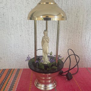 Puede incluir: Una lámpara de lluvia vintage con una cúpula y base doradas. En el interior, una estatua crema de una mujer se alza en medio de vegetación artificial. La lámpara tiene una base negra y un cable de alimentación con enchufe.