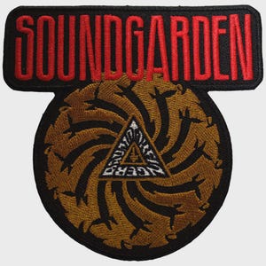 Pode incluir: Um emblema bordado preto com o texto vermelho SOUNDGARDEN numa secção retangular superior. Abaixo, uma secção circular exibe um padrão espiral dourado de silhuetas que rodeia um triângulo central com texto branco e um pequeno ícone.