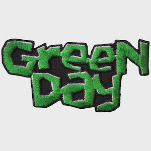Könnte beinhalten: Ein gestickter Aufnäher mit dem Schriftzug Green Day in einer fetten, blockartigen Schriftart. Die Buchstaben sind leuchtend grün mit einem dünnen weißen Rand und auf einem schwarzen Hintergrund platziert, der der unregelmäßigen Form folgt.
