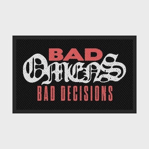 Op de afbeelding: Een rechthoekige zwarte mat met een getextureerd oppervlak met de rode tekst BAD OMENS in een vetgedrukt lettertype, gevolgd door witte gestileerde letters BAD OMENS, en een laatste regel rode tekst BAD DECISIONS in een schreefloos lettertype.
