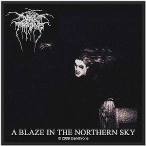 May include: A square black fabric patch featuring a white logo at the top, a central image of a face with white and black makeup, and the text A BLAZE IN THE NORTHERN SKY and 2009 Darkthrone printed in white at the bottom of the design.