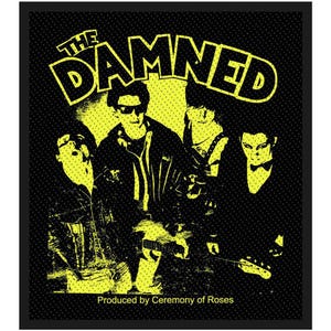Puede incluir: Parche gráfico en negro y amarillo con la banda "The Damned". La imagen muestra una ilustración estilizada de los miembros de la banda. El nombre de la banda está en una fuente amarilla en negrita en la parte superior. El texto "Produced by Ceremony of Roses" está en la parte inferior.