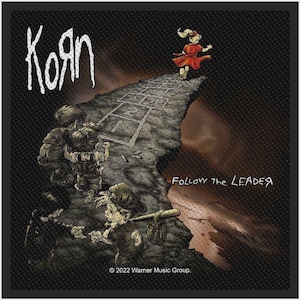 Op de afbeelding: Een zwarte stoffen patch met het Korn-logo en de tekst Follow the Leader. De afbeelding toont kinderen op een klif met een hinkelspel, een kind in een rode jurk en een ander met een honkbalknuppel. Bevat de tekst 2022 Warner Music Group.