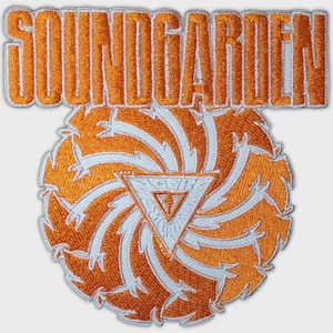 May include: An embroidered patch featuring the word SOUNDGARDEN in orange block letters above a circular design. The circle contains a white triangular emblem with a central torch icon, surrounded by orange and white swirling, flame-like patterns.