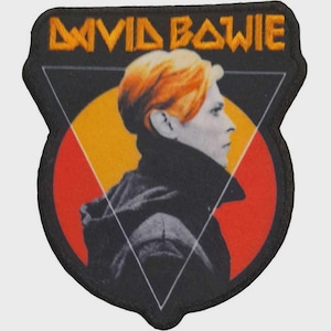 Puede incluir: Un parche bordado en forma de escudo con un retrato de perfil de una persona con cabello naranja y chaqueta negra. El fondo incluye un círculo rojo y un triángulo blanco. El texto DAVID BOWIE está bordado en amarillo en la parte superior.