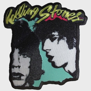 Può includere: Una toppa ricamata rettangolare con la scritta Rolling Stones in corsivo giallo nella parte superiore. Sotto il testo ci sono due ritratti in bianco e nero di persone con capelli scuri su uno sfondo verde acqua chiaro con bordo nero.