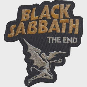 Puede incluir: Un parche bordado negro con el texto BLACK SABBATH en hilo dorado y THE END en hilo plateado. Debajo del texto hay una figura alada bordada en plata en una pose dinámica, todo sobre un fondo de tela negra con un borde definido.