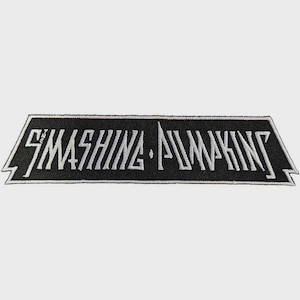 May include: A rectangular embroidered patch with a black background and white border. The text SMASHING PUMPKINS is stitched in a stylized white font across the center. The patch features pointed, notched edges on both the left and right sides.