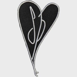 May include: A black heart-shaped embroidered patch featuring a white stitched border and a white stylized script monogram in the center. The design consists of a large heart outline with a cursive letter pattern integrated into the black fabric surface.