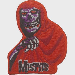 Può includere: Una toppa ricamata raffigurante una figura con cappuccio rosso e volto scheletrico nei toni del viola, bianco e nero. La figura ha occhi rossi e una mano scheletrica. La parola Misfits è ricamata in bianco con un contorno nero sul fondo.