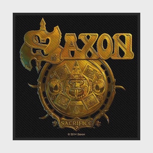 Könnte beinhalten: Ein quadratischer Aufnäher mit einem goldfarbenen, metallischen Design auf schwarzem Hintergrund. Das Wort SAXON ist in großen, stilisierten Buchstaben über einem kreisförmigen Emblem abgebildet, das einem aztekischen Kalender ähnelt.